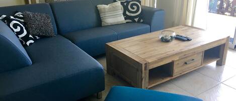 Een woonkamer met een blauwe bank, een houten salontafel en een eethoek met een tafel en stoelen.