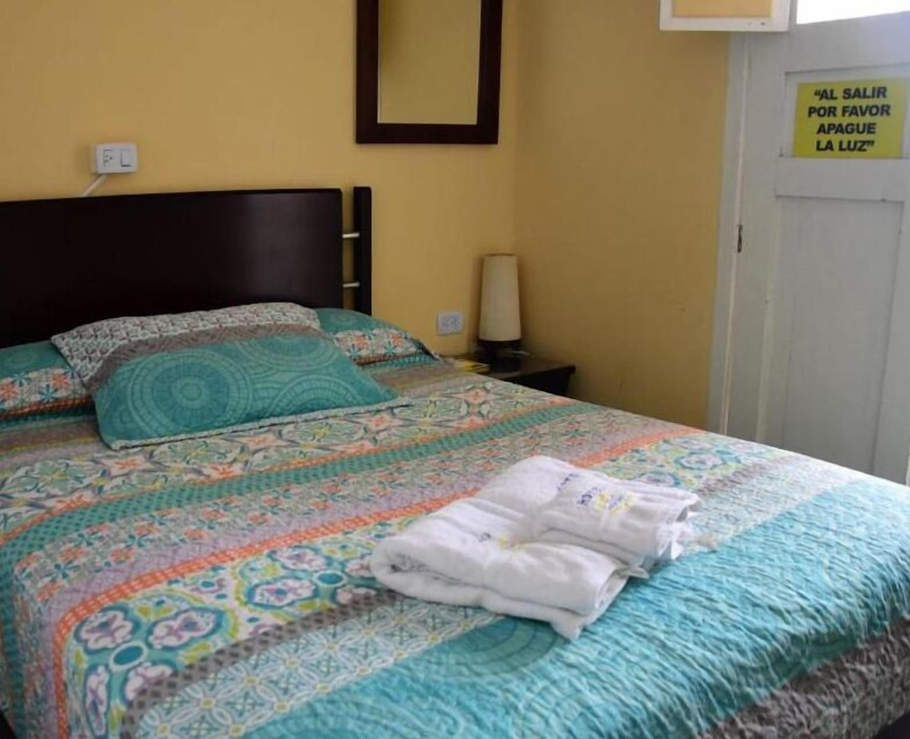 Una cama bien hecha con ropa de cama estampada y toallas dobladas, una lámpara de noche y una puerta con un cartel en español.