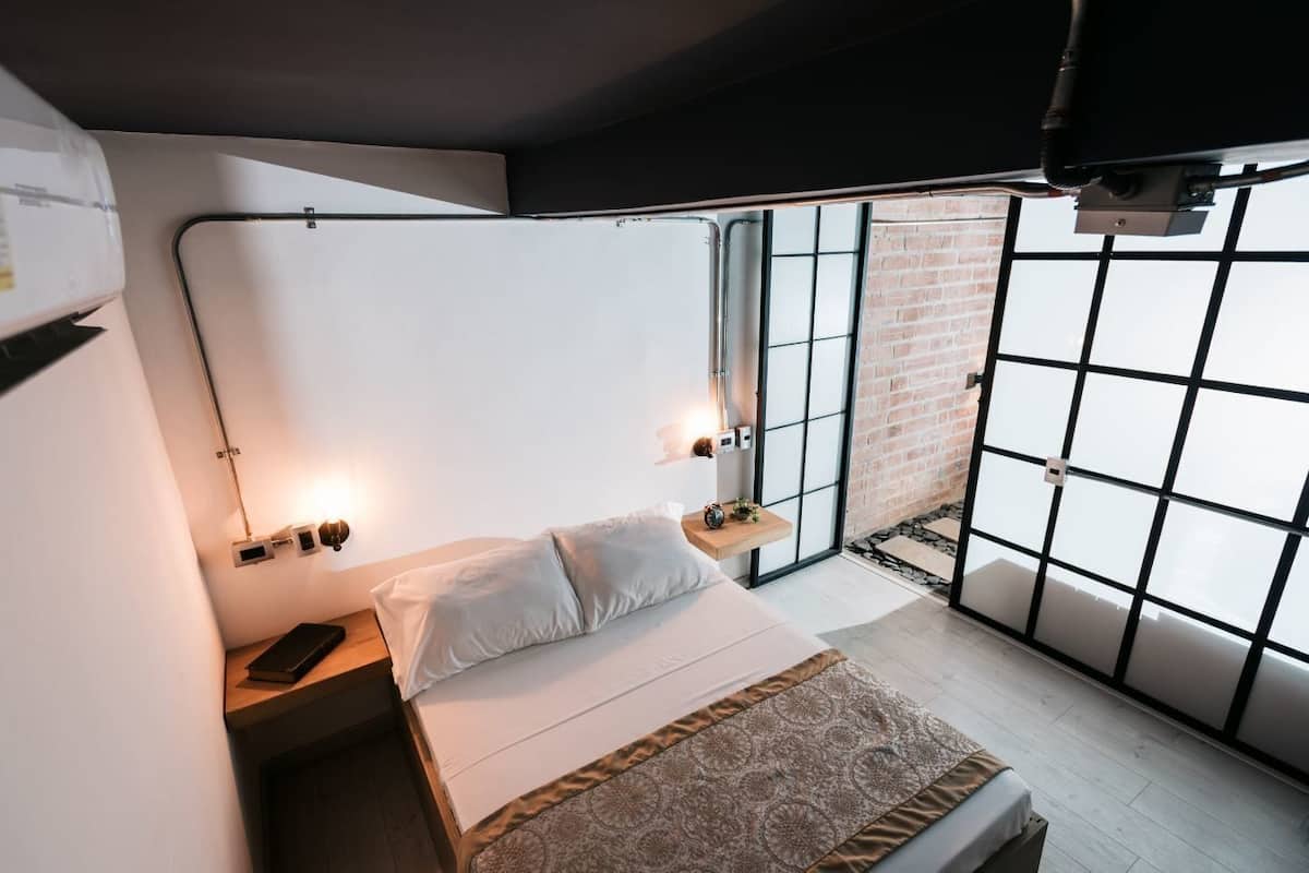 Un dormitorio moderno con cama, mesitas de noche, lámpara colgante y ventana con vistas a un muro de ladrillo.