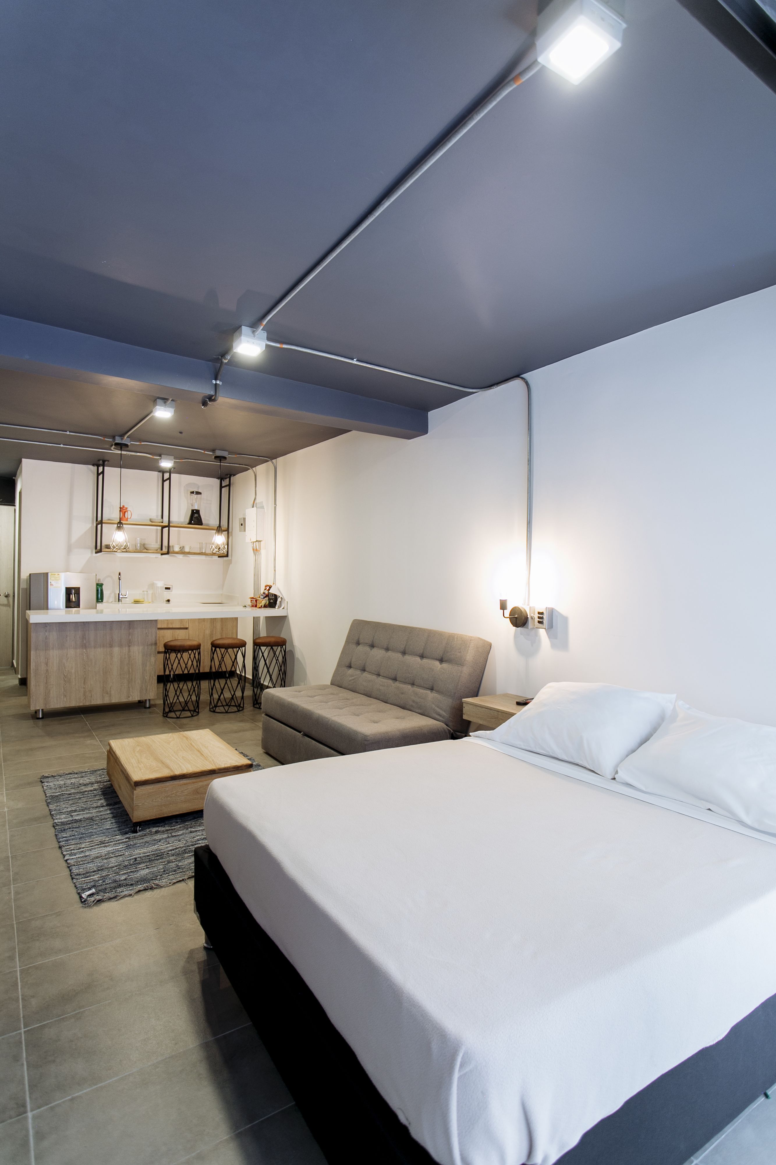 A modern bedroom with a large bed, a sofa, a small kitchen area with stools, and a dining table.