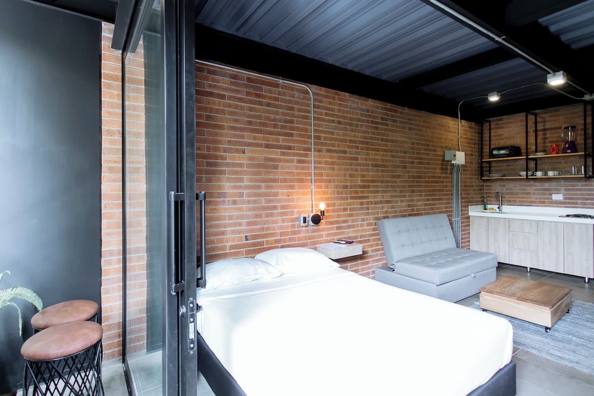 Una habitación de hotel moderna con pared de ladrillo, cama, sofá gris, una mesita pequeña y una kitchenette.