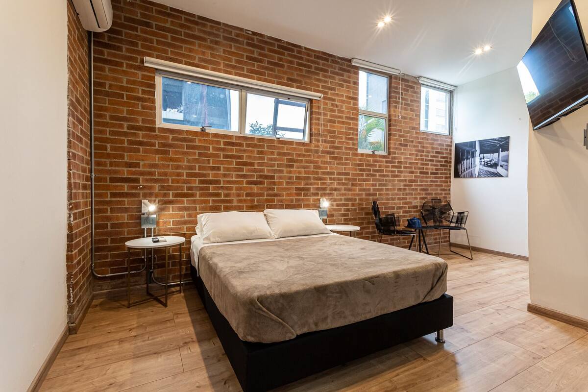 Un dormitorio con pared de ladrillo, una cama, dos mesitas de noche, una silla, una mesita redonda pequeña y un televisor montado en la pared.