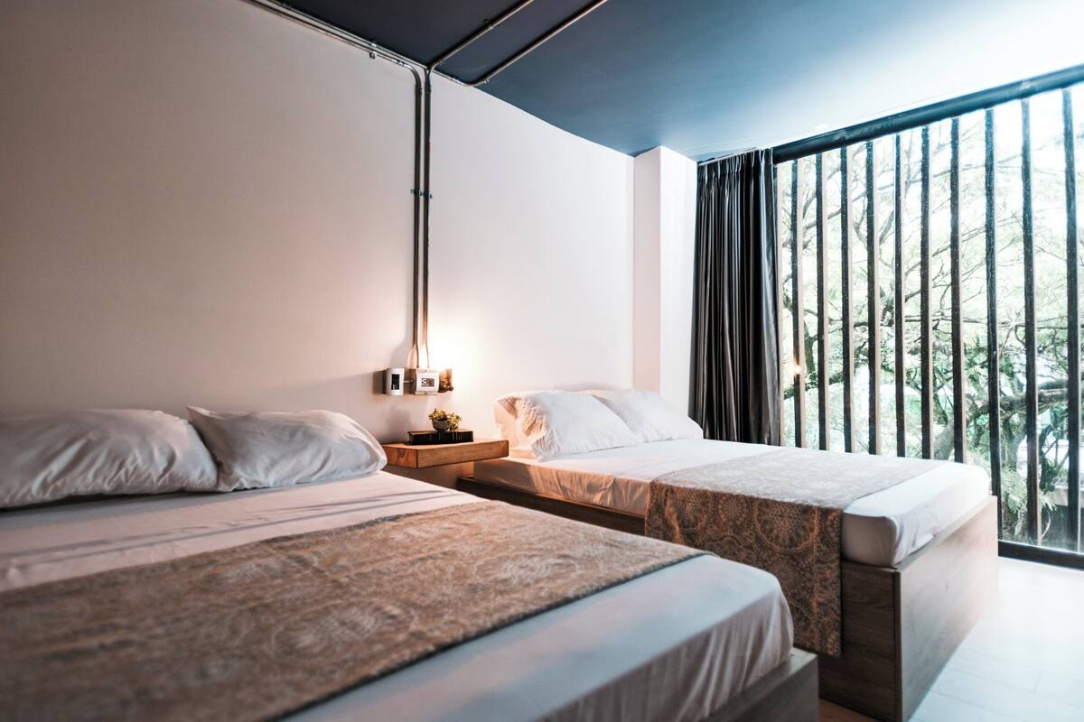 Un dormitorio moderno con dos camas, una mesita de noche y un ventanal con vistas a árboles.