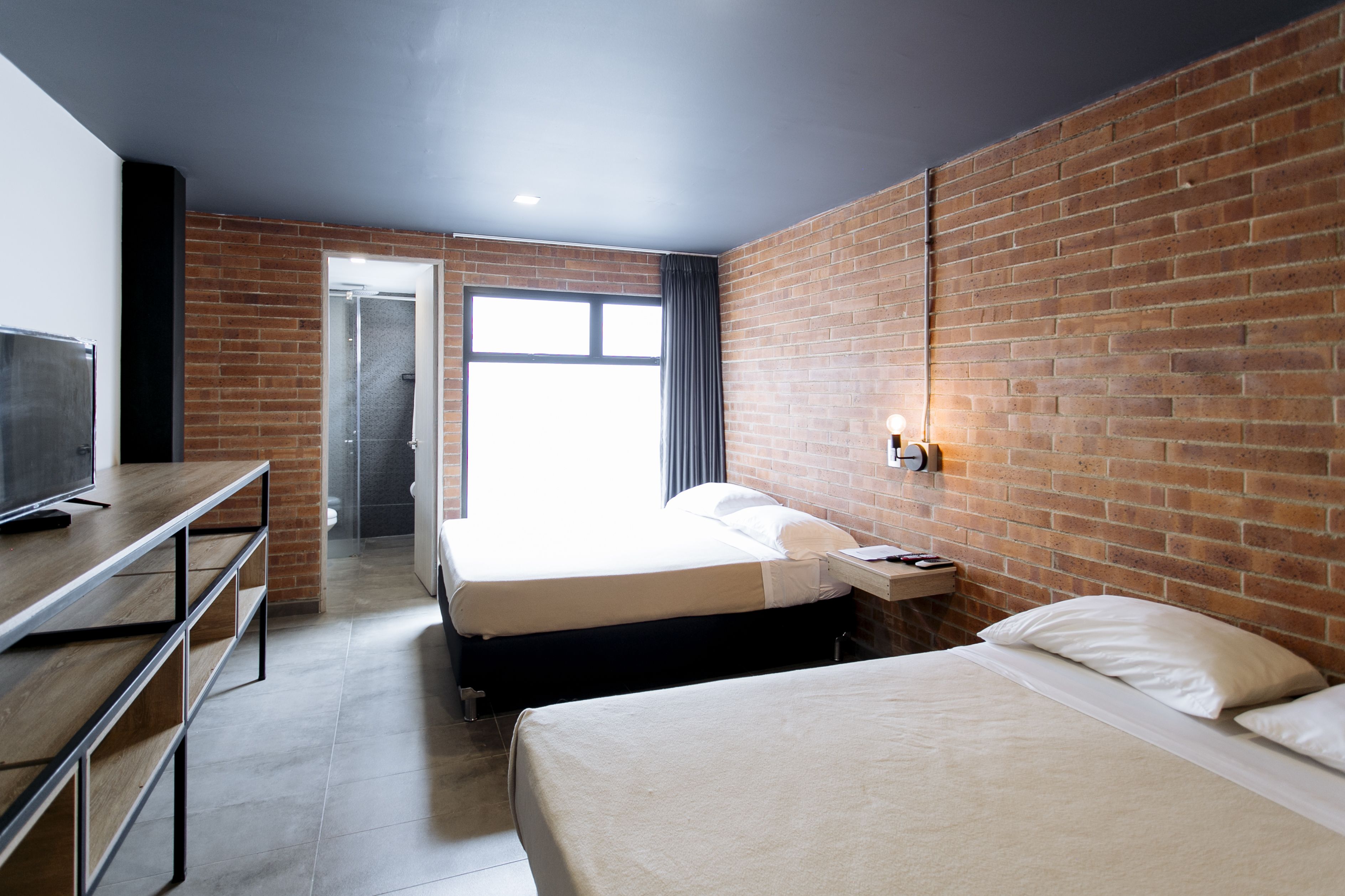 A hotel room with a brick wall, a bed, a desk with a TV, and a window.