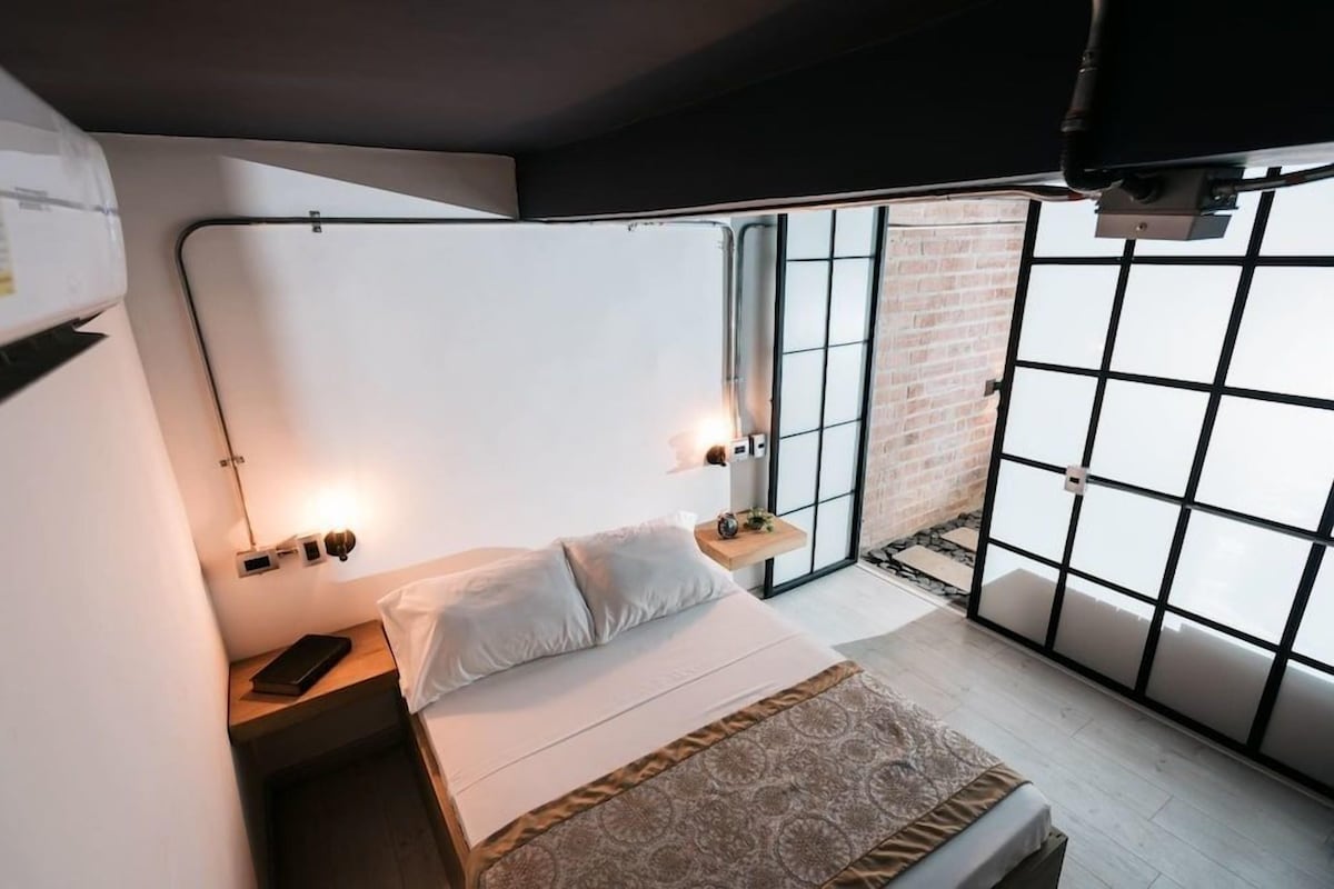 Une chambre à coucher avec un lit, une table de chevet, une lampe et une vue sur un mur de briques, visible par une fenêtre.
