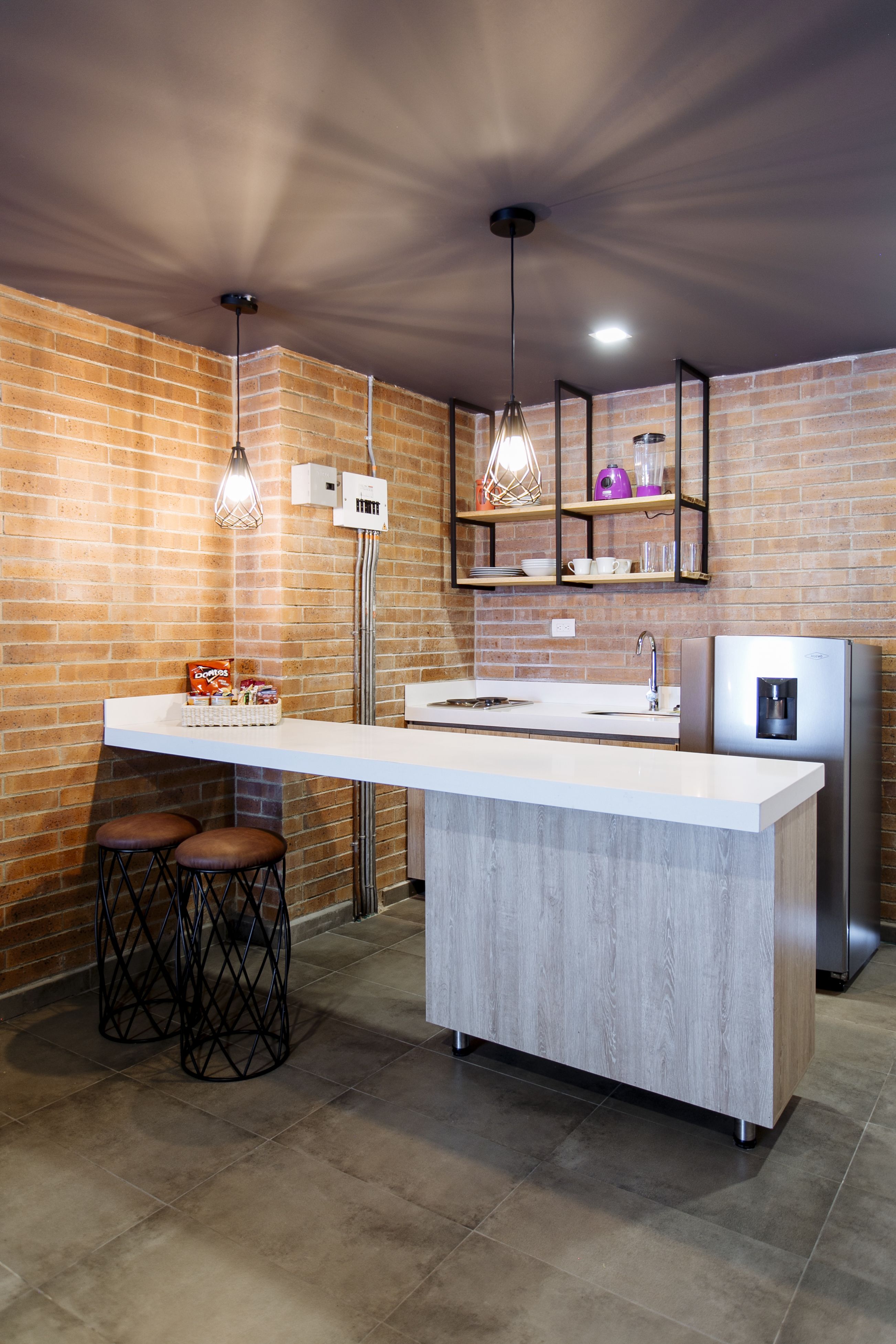 A modern kitchen with a central island, bar stools, a refrigerator, pendant lights, and a brick wall.