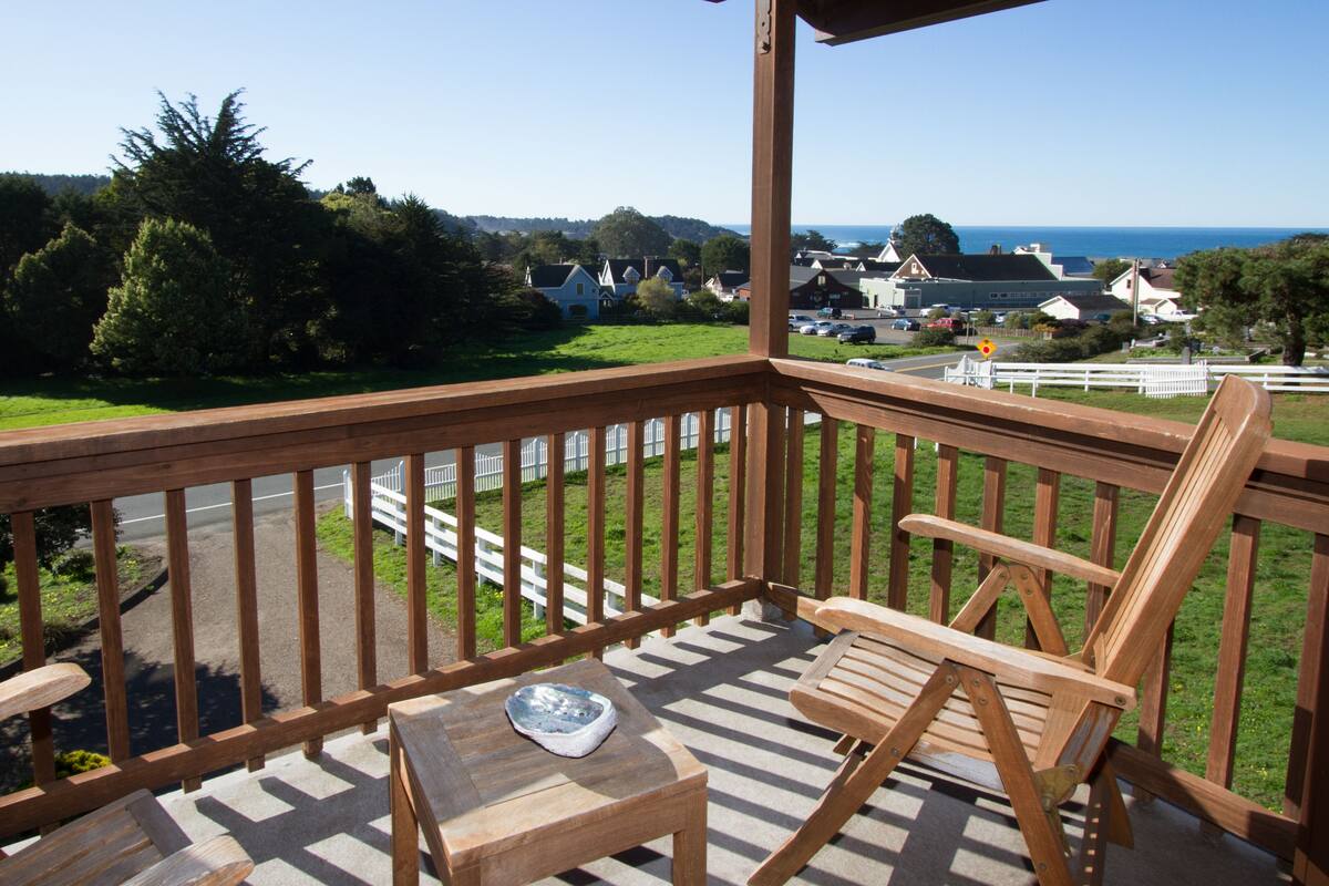 A wooden deck with a table and chairs, overlooking a road and houses with a view of the sea.