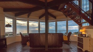 A spacious room with a large window overlooking the ocean, a wooden ceiling, and a staircase leading to an upper level. - Sweetwater Vacation Rentals (Mendocino)