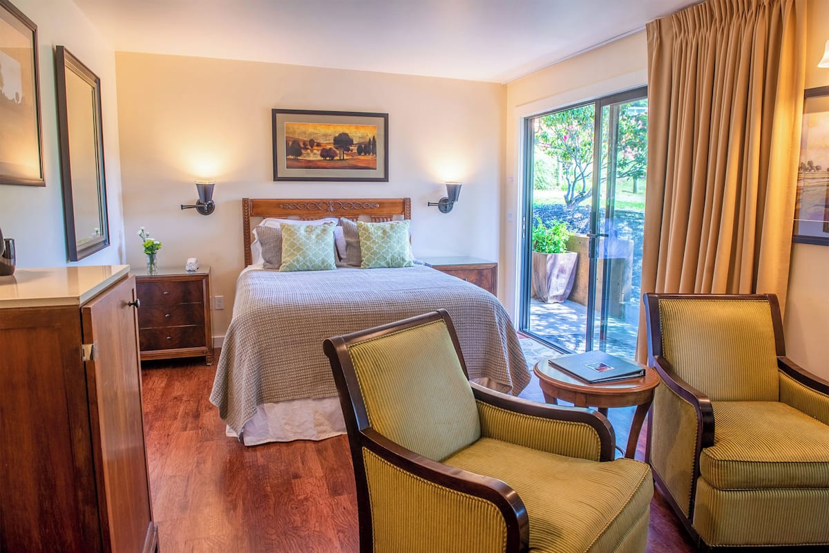 A hotel room with a bed, two chairs, a nightstand, a lamp, and a painting on the wall.