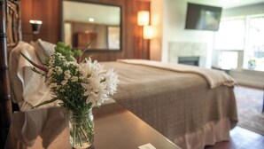 A hotel room with a bed, a bedside table with a vase of flowers, a lamp, and a television mounted on the wall. - Inn at Buckhorn Cove (Little River)