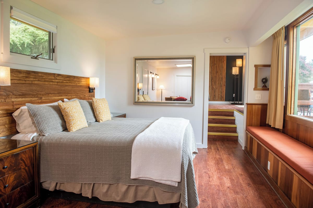A bedroom with a large bed, a wooden headboard, a mirror, a bench, and a view of the outdoors.