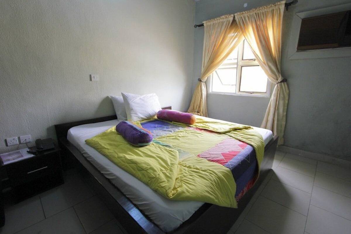 A single bed with a yellow quilt, a white pillow, and a purple pillow. There is a nightstand with a lamp and a power outlet. The room has a window with beige curtains and a wall-mounted air conditioning unit.