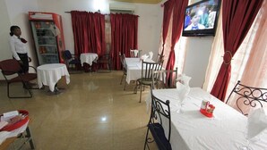 A hotel room with a TV, red curtains, a small table with a red tray, and a person standing by a bookshelf. - Sylva Link Hotel (Lagos)
