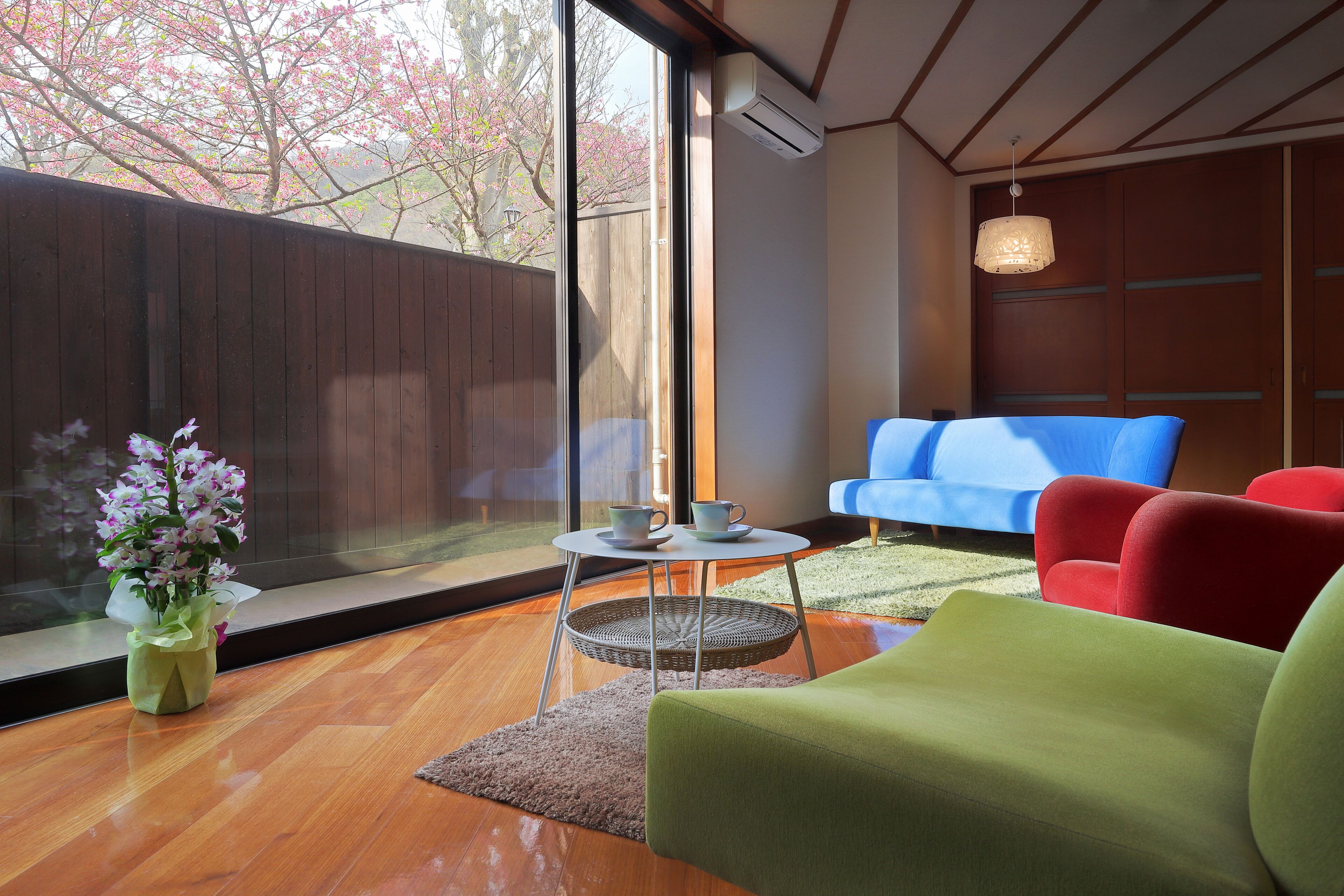 A modern living room with a large window, a blue sofa, a red armchair, and a green sofa.