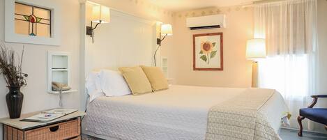 A neatly arranged bedroom with a bed, bedside lamps, a wall-mounted telephone, a framed picture, and a small table with a vase and a basket.