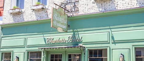 Un bâtiment en briques vertes sur lequel on peut lire l’inscription « Charlotte Hotel Restaurant ». Des plantes en pot sont disposées à l’extérieur et un banc se trouve sur la droite.