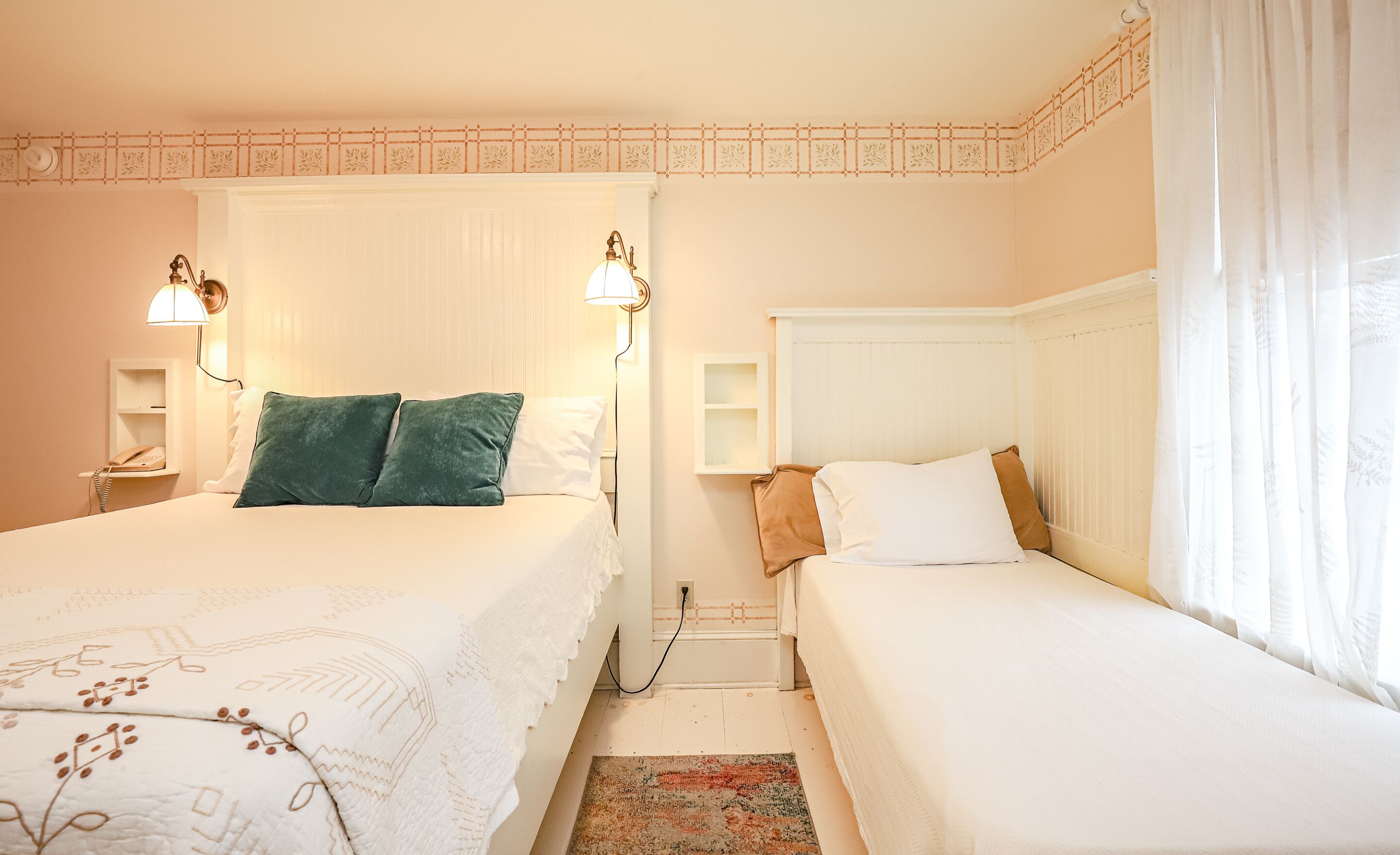 Habitación de hotel con dos camas, una mesita de noche con lámpara, un panel decorativo en la pared y una ventana con cortinas traslúcidas.