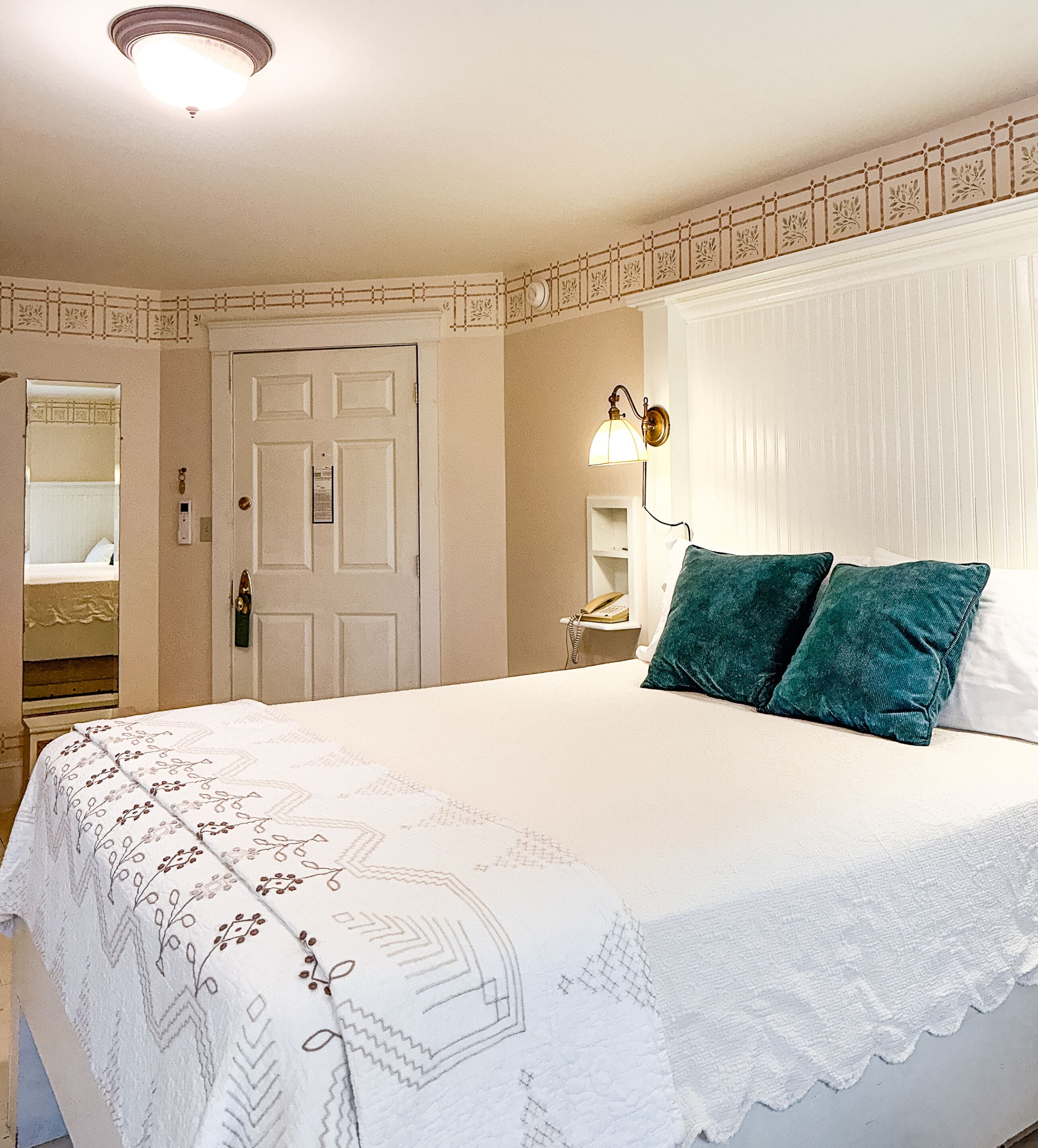 Une chambre à coucher avec un grand lit, une table de chevet, une lampe fixée au mur et une porte donnant sur une autre pièce.