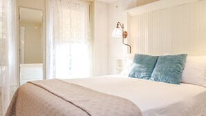 Uma cama bem arrumada com um edredom bege e duas almofadas azuis, um abajur de cabeceira e uma janela com cortinas leves.