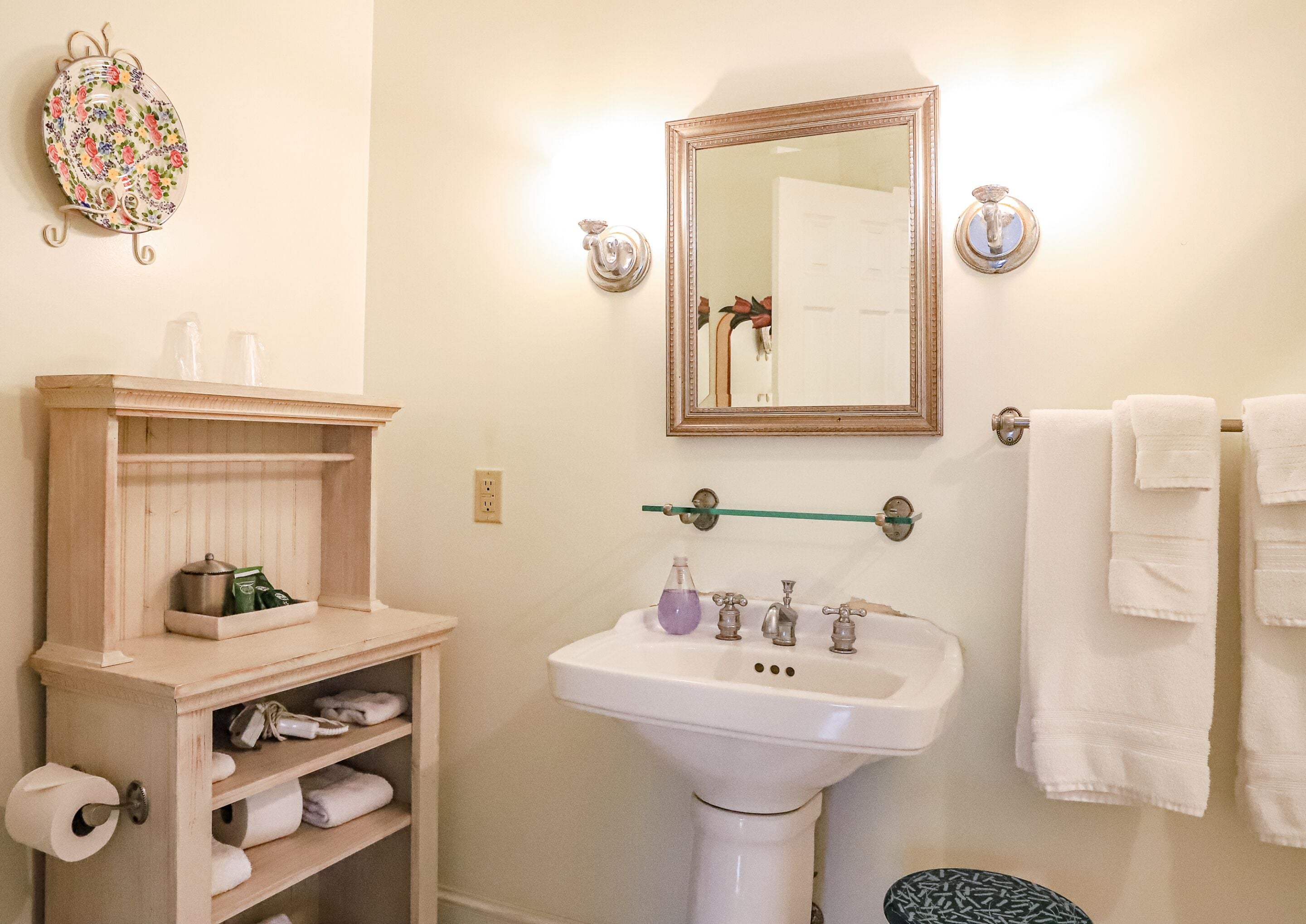 A bathroom with a wooden shelf holding toiletries, a white sink with three faucets, and a mirror above it.
