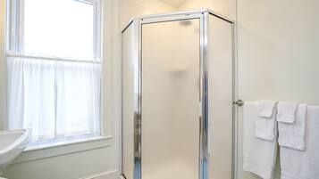 A bathroom with a glass shower enclosure, a window with sheer curtains, and a sink with a white countertop.