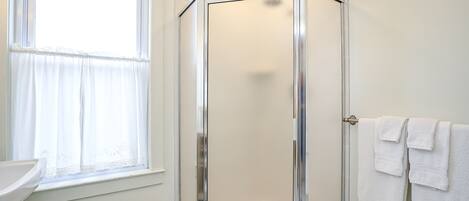 A bathroom with a glass shower enclosure, a window with sheer curtains, and a sink with a white countertop.