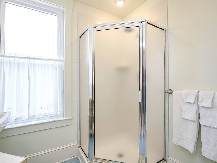 A bathroom with a glass shower enclosure, a window with sheer curtains, and a sink with a white countertop.
