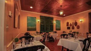 Restaurante com várias mesas preparadas para refeições, com decoração clássica, cadeiras de madeira e toalhas de mesa brancas.
