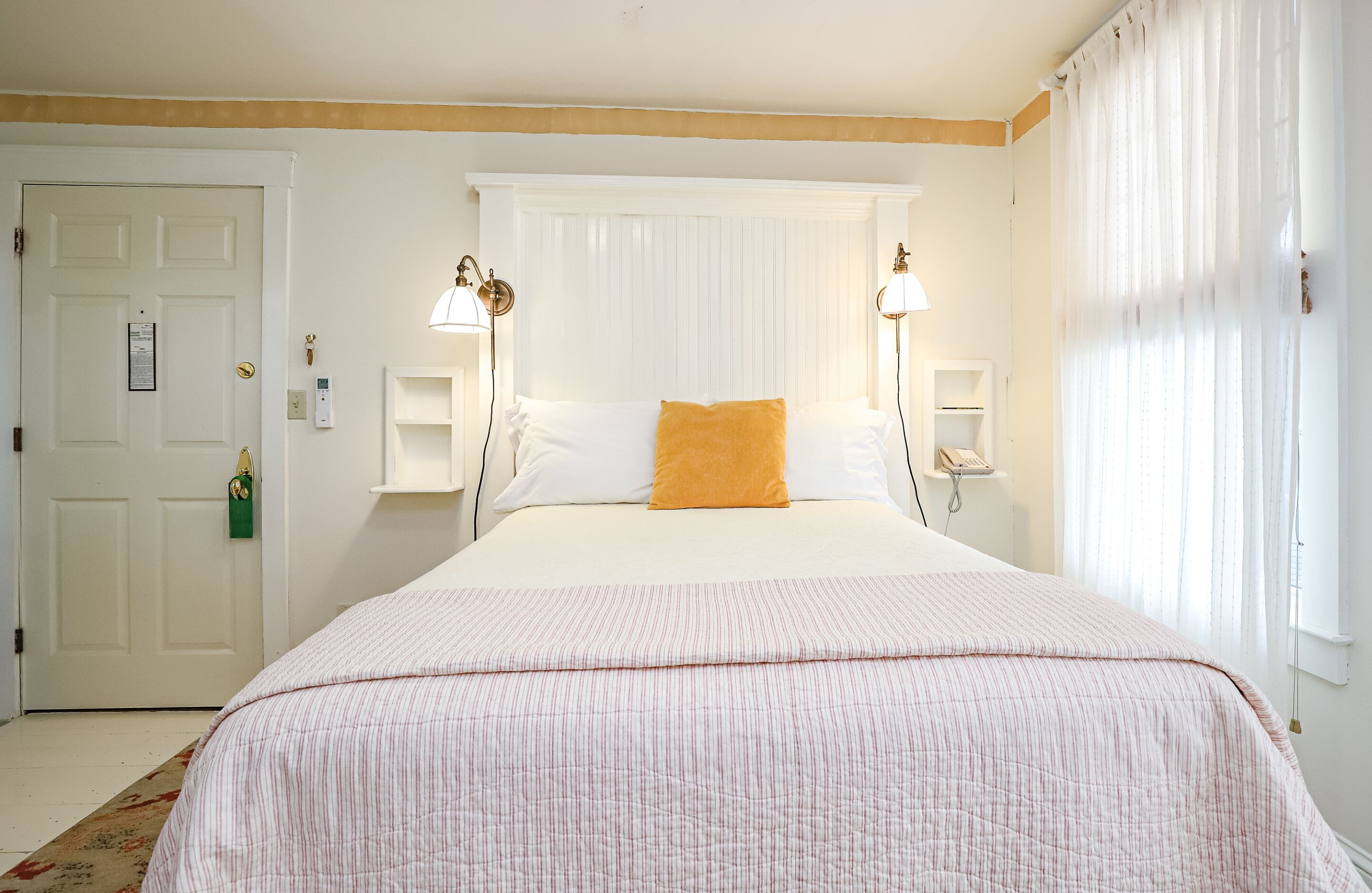 A neatly arranged bedroom with a large bed, two bedside lamps, a window with sheer curtains, and a door with a green keychain.