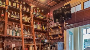 Um bar com prateleiras de madeira repletas de garrafas de bebidas, uma televisão presa na parede e um amplo janelão que ilumina naturalmente o espaço.