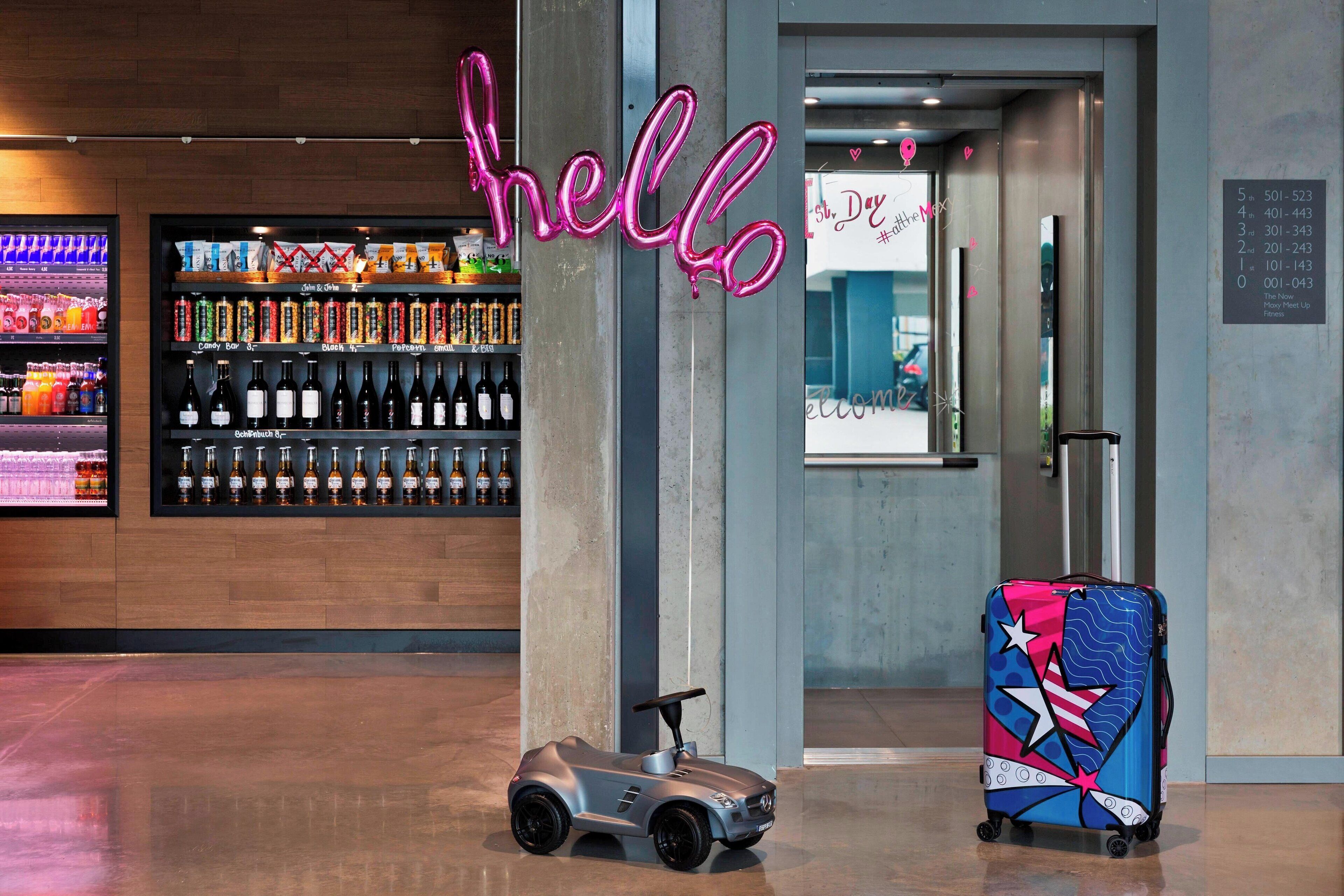 A modern hotel lobby with a neon 'hello' sign, a bar area with various beverages, and a colorful suitcase.