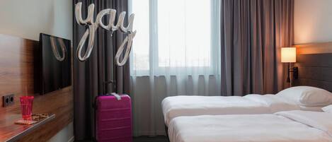 Quarto de hotel com duas camas, uma janela grande com cortinas, uma mala rosa, uma TV na parede e uma decoração com balão escrito 'yay'.