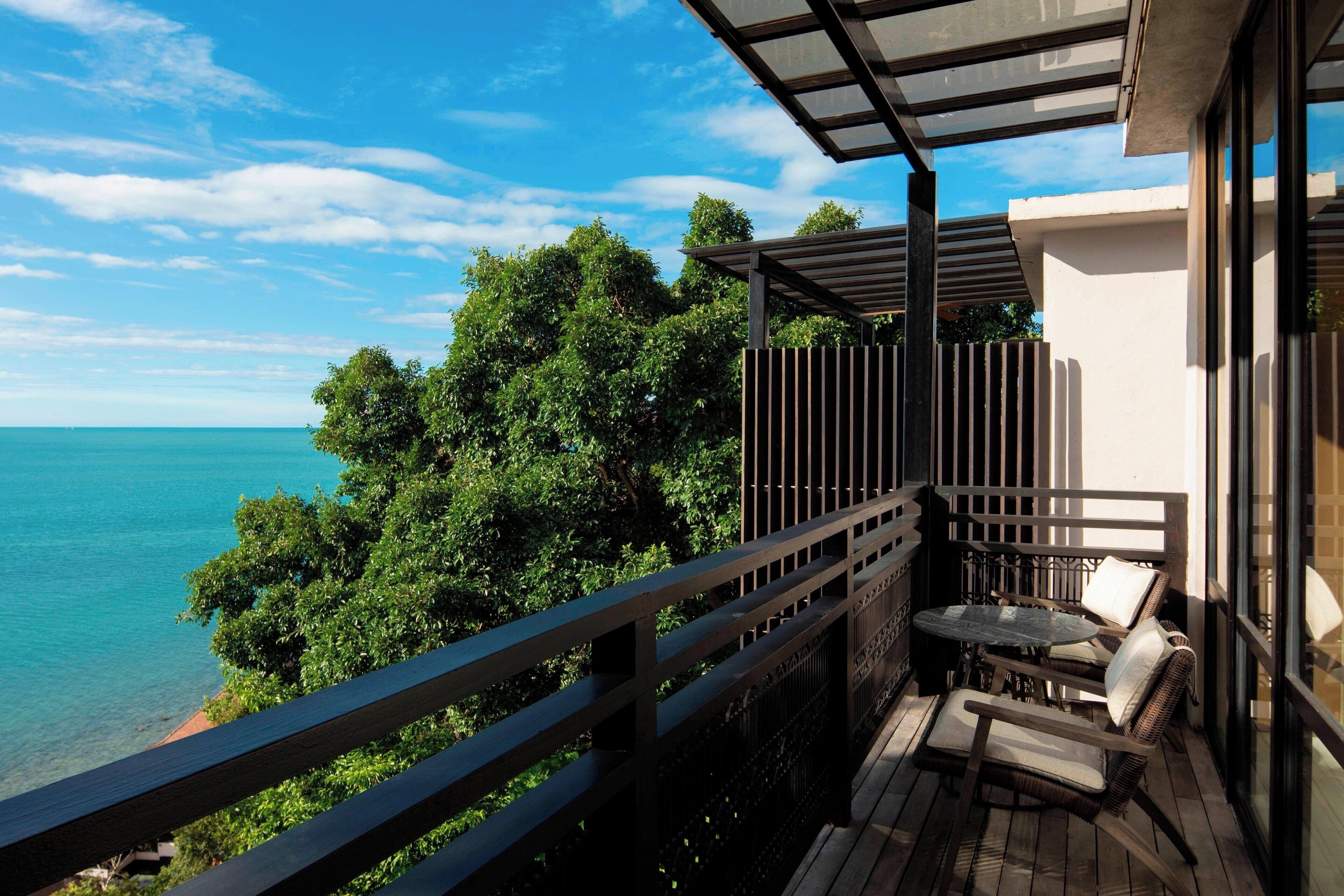 Un balcon avec vue sur l’océan et les arbres, une table avec des chaises et une véranda fermée.