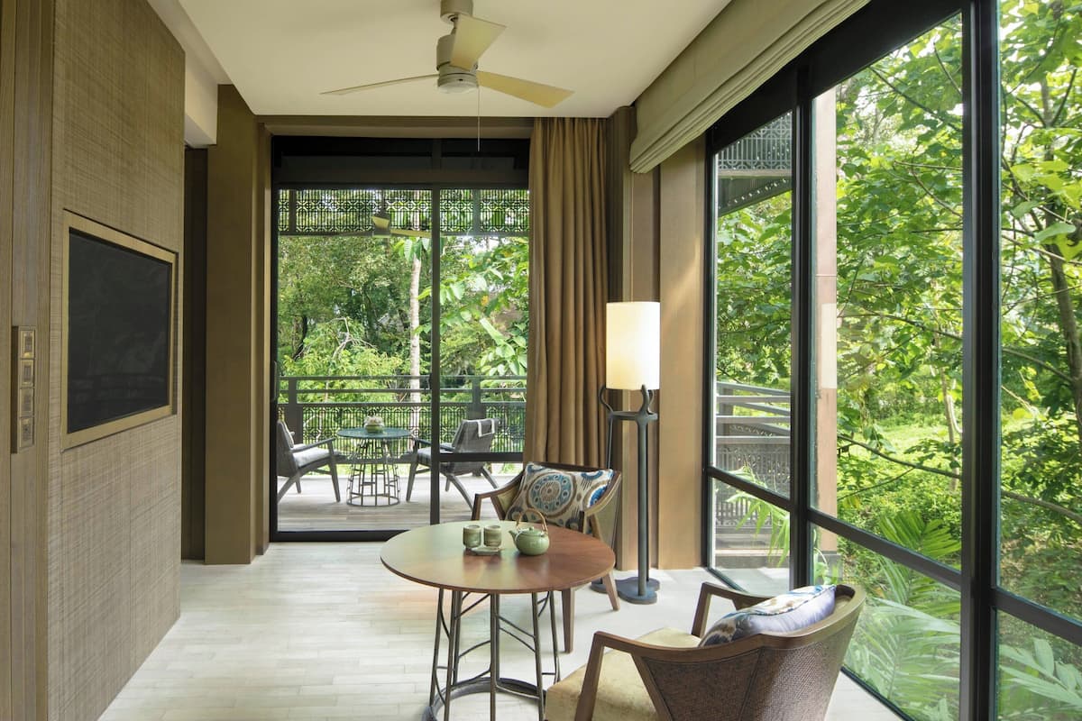 A room with a large window, a ceiling fan, a table with chairs, and a TV on the wall.
