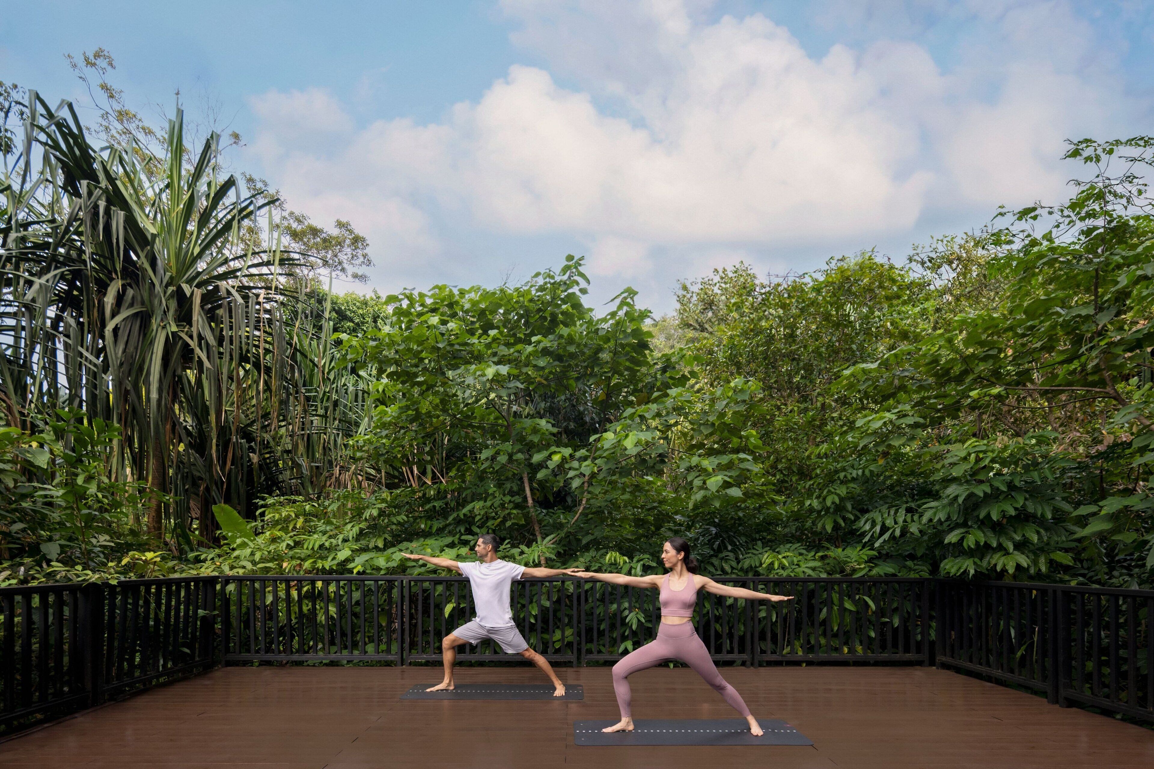 Dos personas practicando yoga en una terraza al aire libre con piso de madera, rodeado de abundante vegetación y un cielo despejado.
