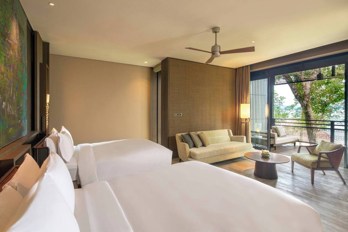A hotel room with two beds, a sofa, a coffee table, and a balcony with outdoor furniture.