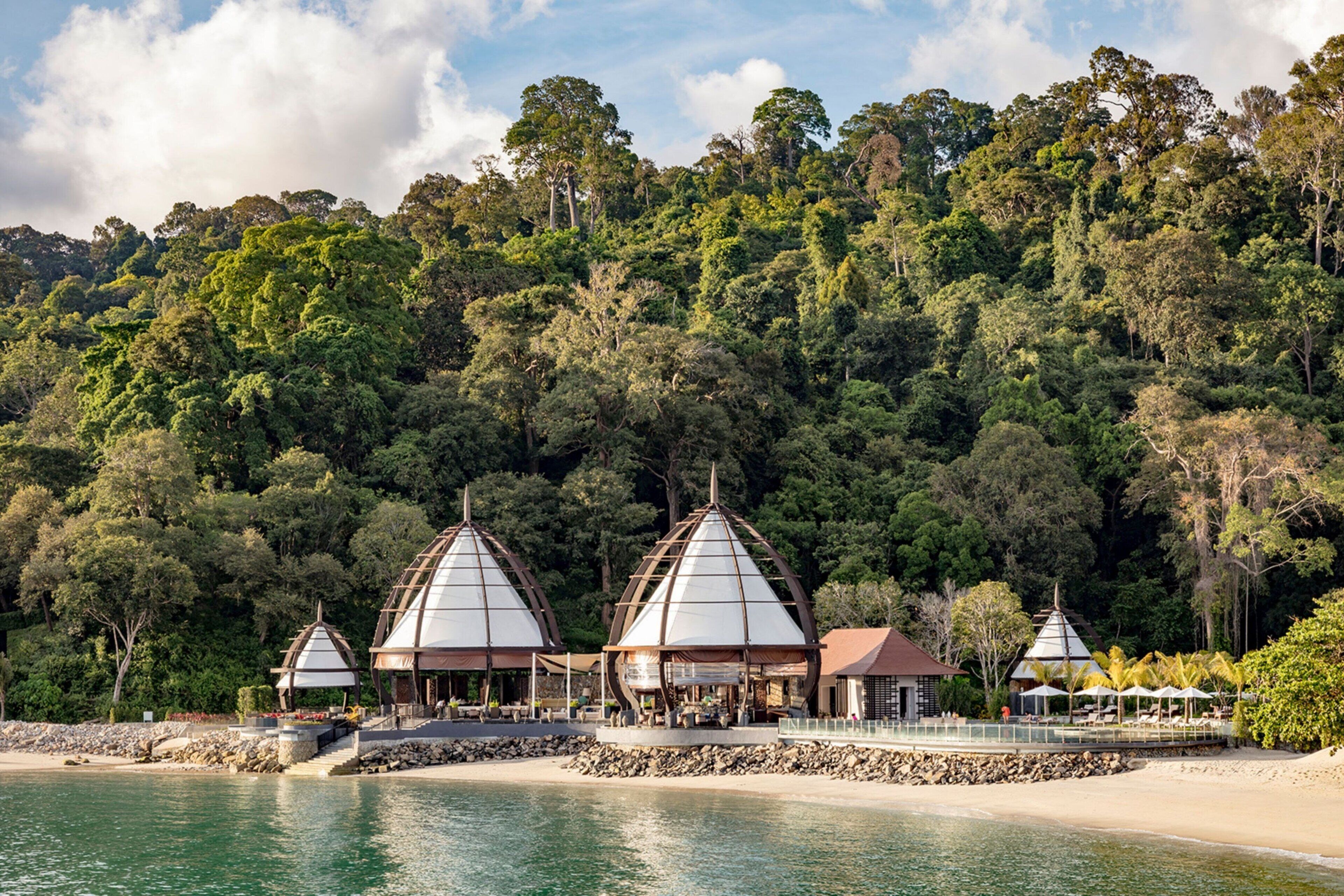 Une station balnéaire dotée de cabanes sur la plage, d’une piscine et bordée d’une forêt.
