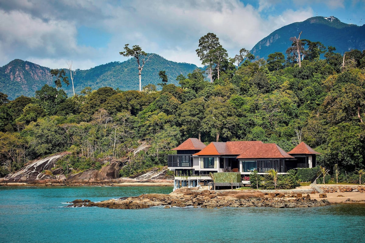 A coastal property with two buildings featuring red roofs, situated on a rocky shoreline with a backdrop of lush green hills and a mountain.