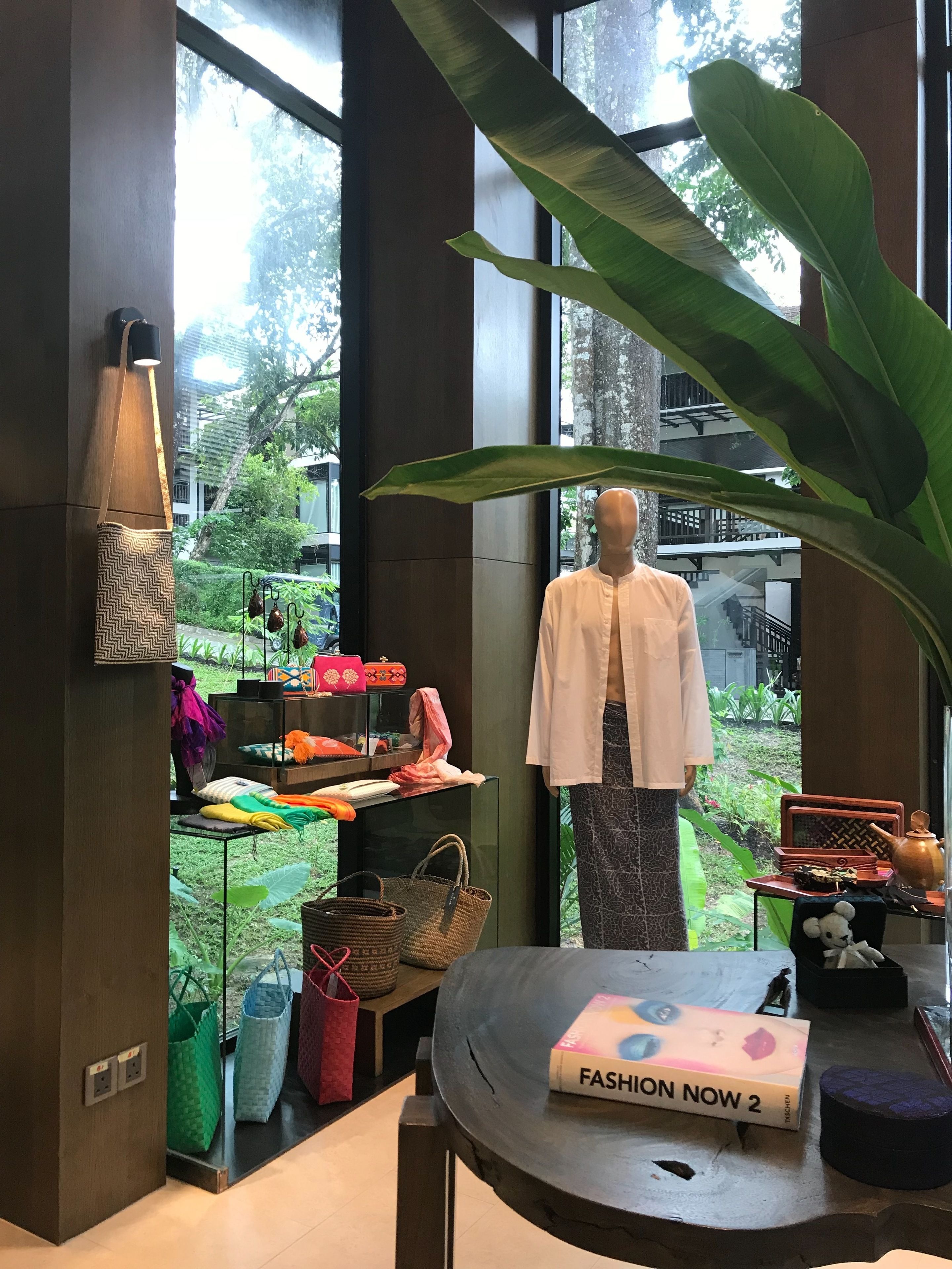 Un maniquí con camisa blanca y pantalones estampados, una mesa con una caja que dice 'Fashion Now 2', y varias bolsas y tejidos en estantes.