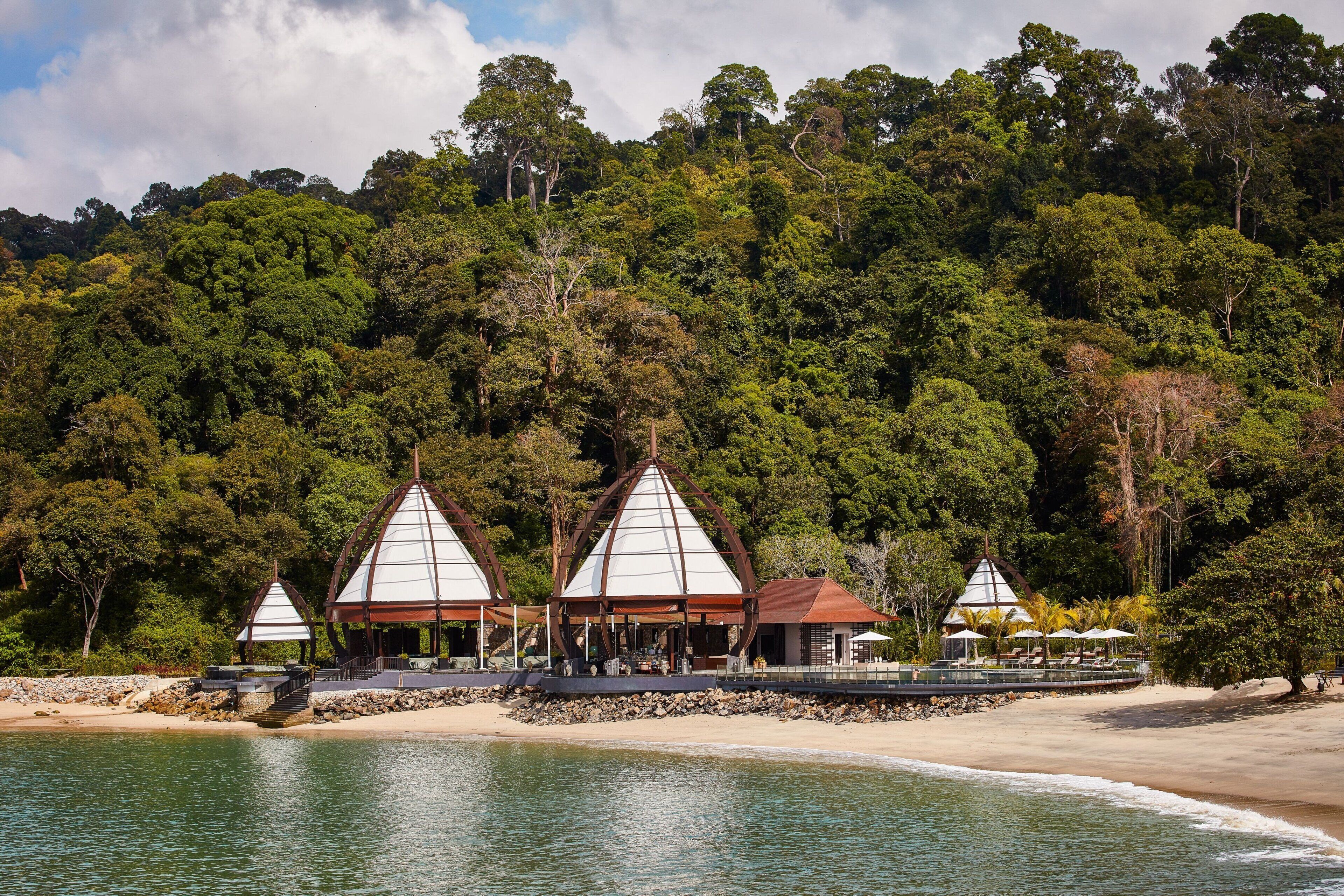 A beachside resort with three conical-roofed structures, a sandy beach, and a forested hillside.