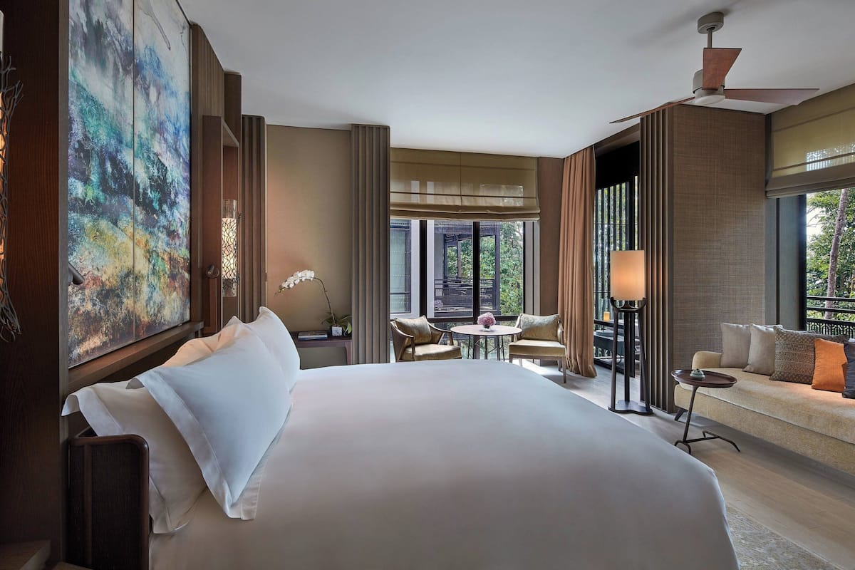 A hotel room with a large bed, a sofa, a small table, and a balcony with a view of the outdoors.