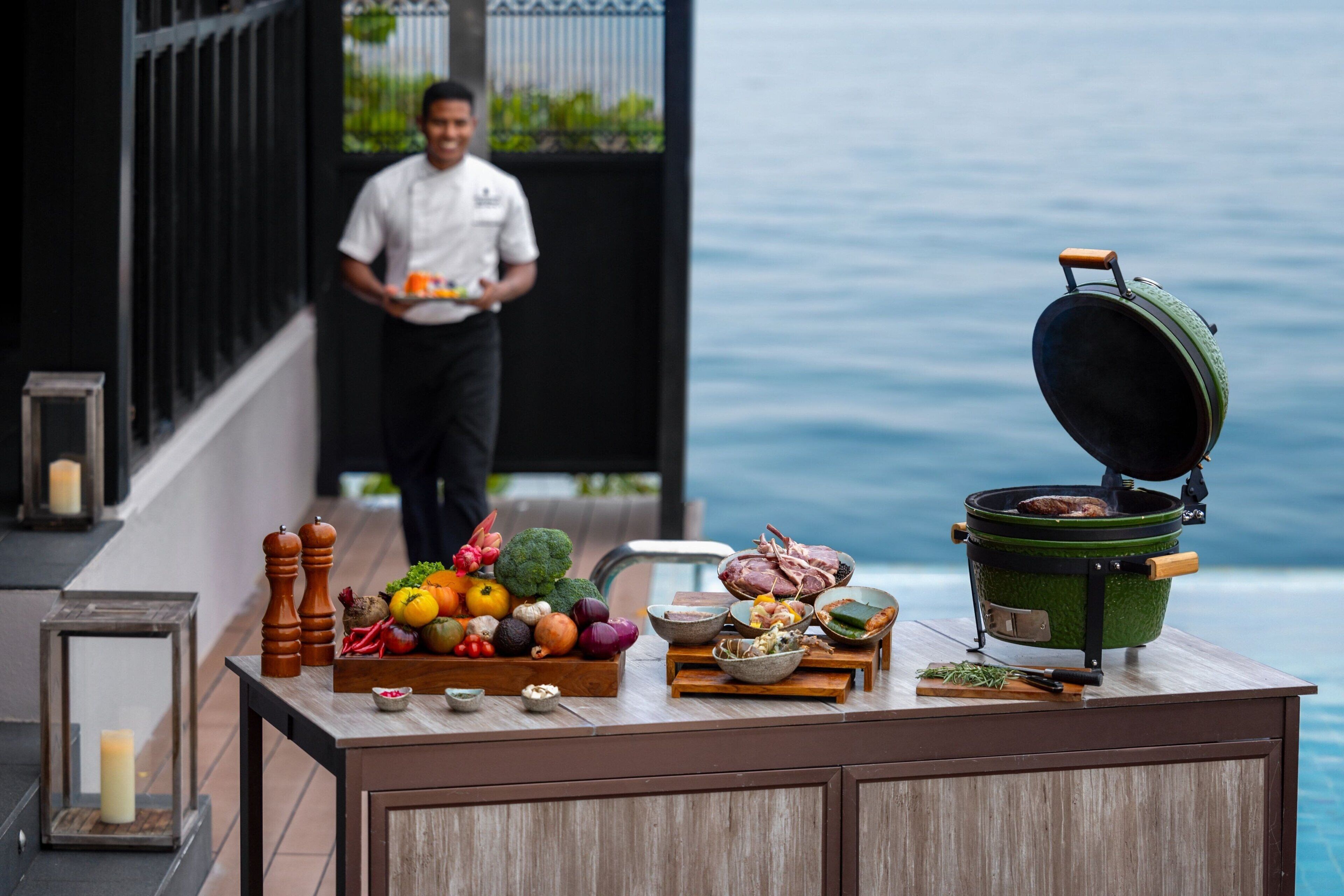 A kitchen counter with a variety of fresh produce, a large pot, and a person carrying a tray.