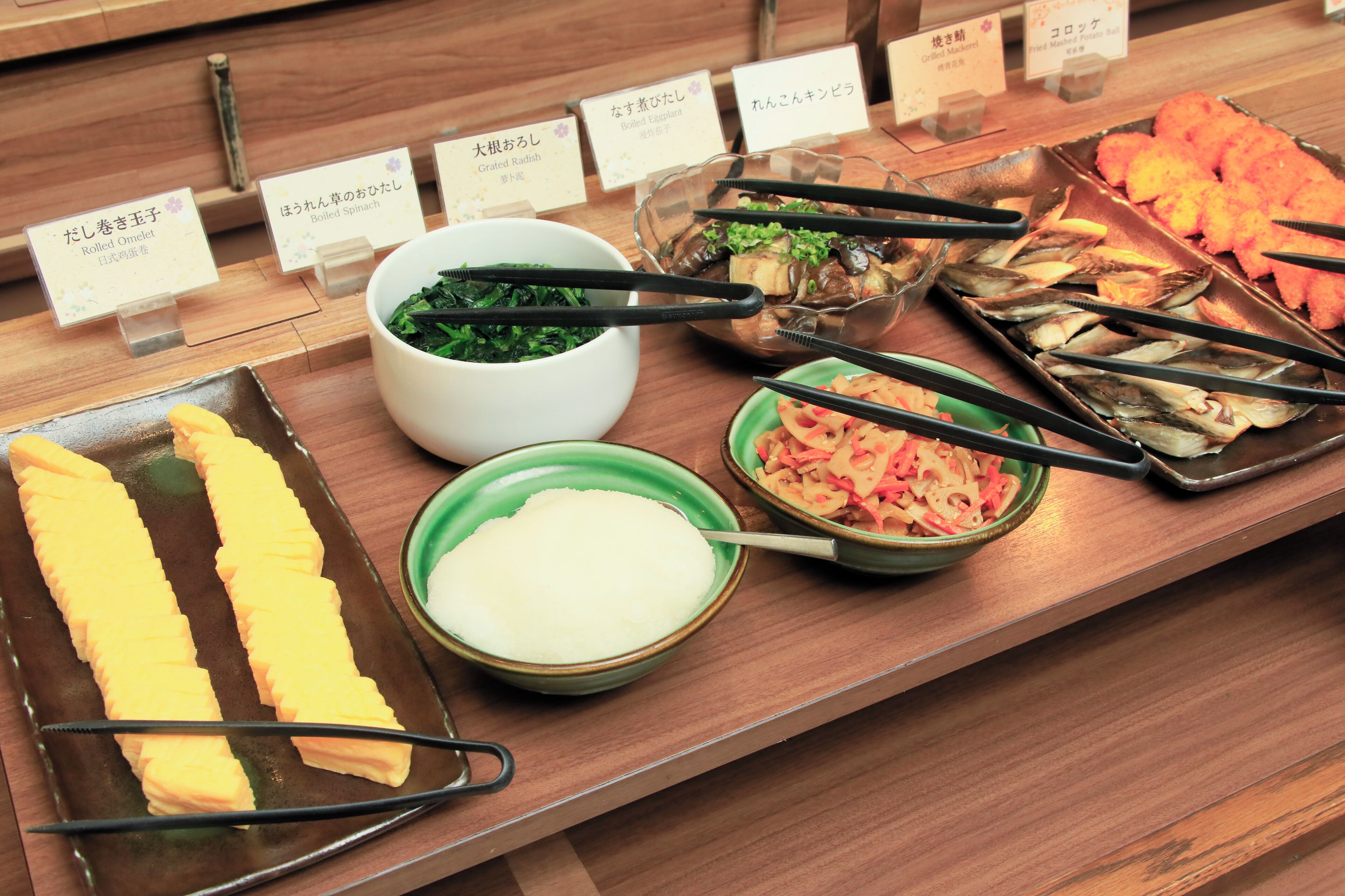 A buffet with various dishes including rolled omelet, boiled spinach, and fish.