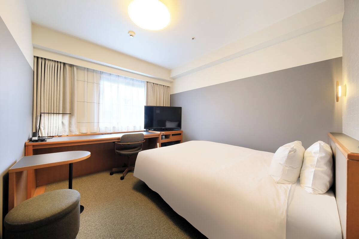 A hotel room with a bed, a desk with a chair, a television, and a window with curtains.