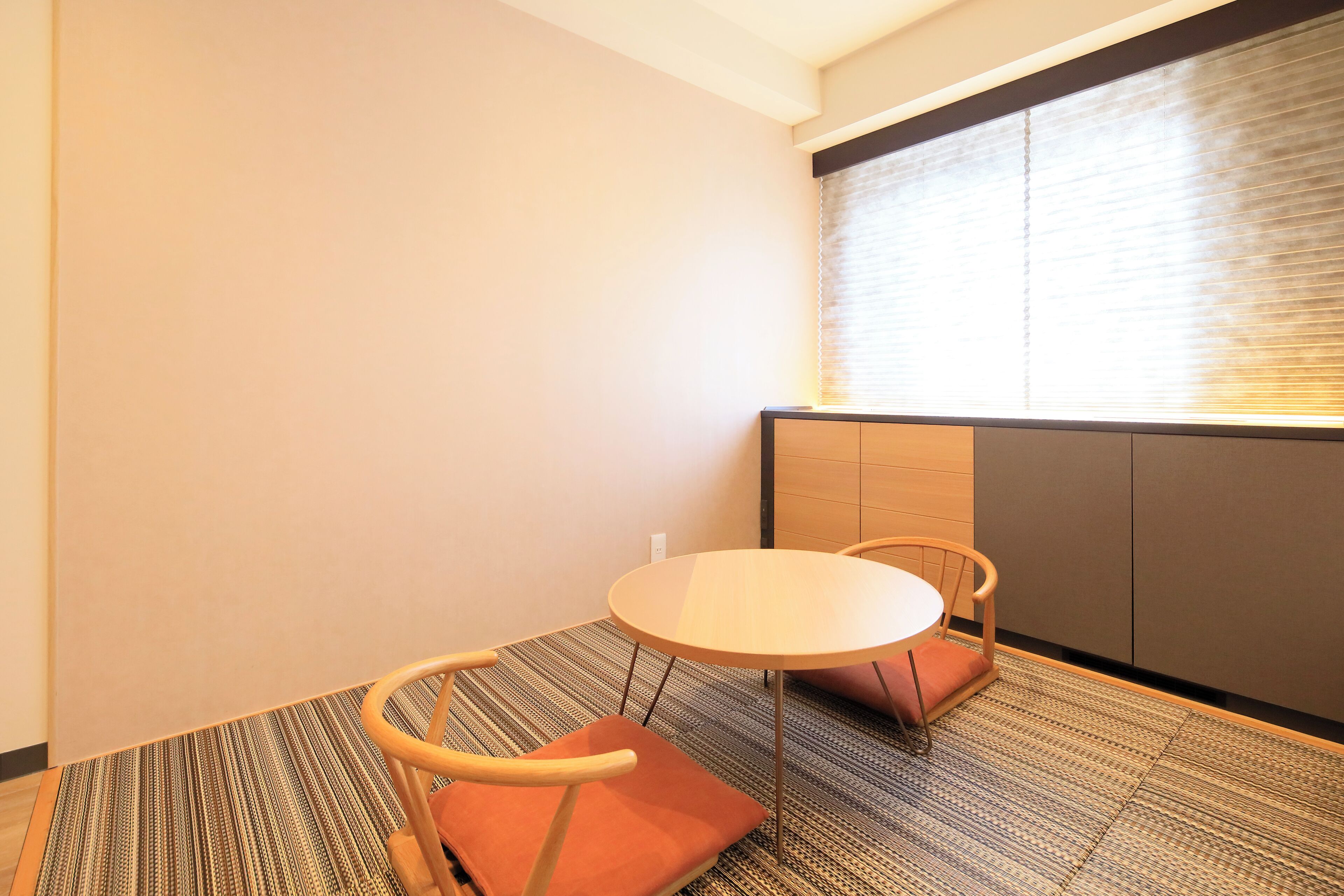 Una habitación pequeña y moderna con una mesa redonda y dos sillas, una ventana con persianas y suelo alfombrado.