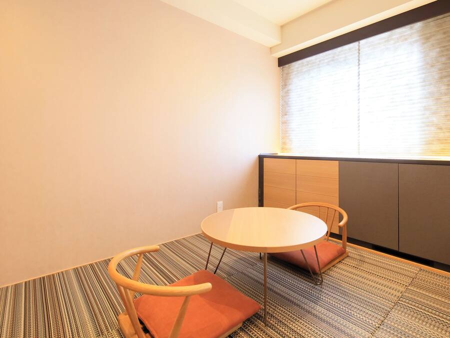 Um quarto pequeno e moderno, com uma mesa redonda e duas cadeiras, uma janela com persianas e chão coberto de carpete.