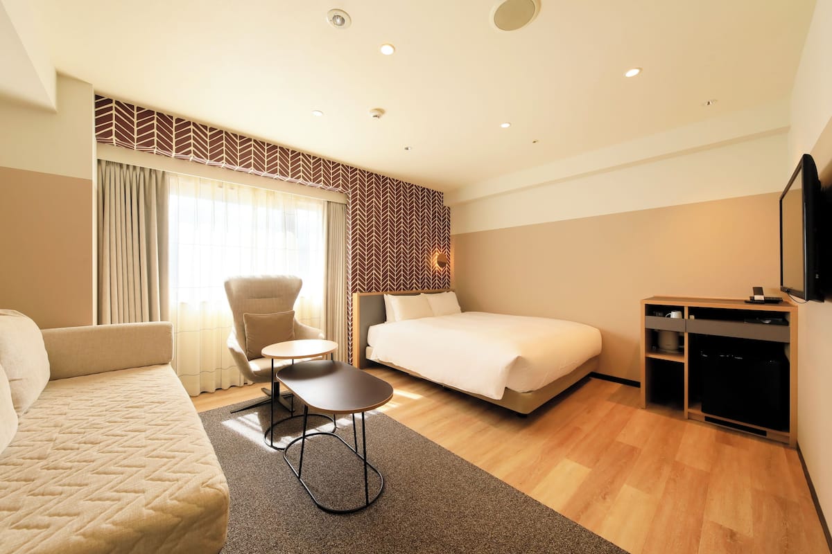 A hotel room with a bed, a sofa, a small table, a chair, a TV, and a wardrobe.