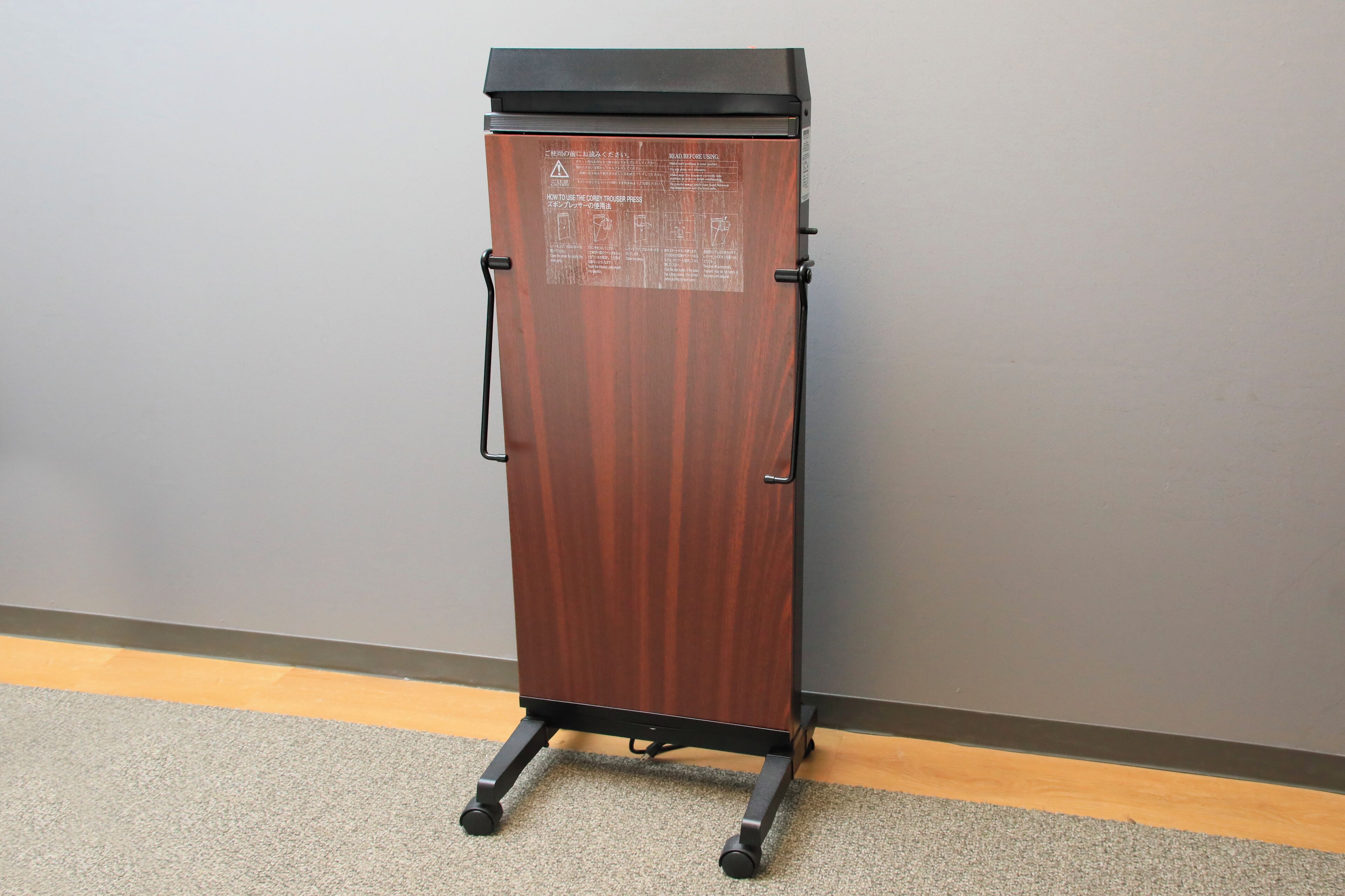 A tall, narrow wooden cabinet with a black top and metal handles, standing on a carpeted floor against a grey wall.