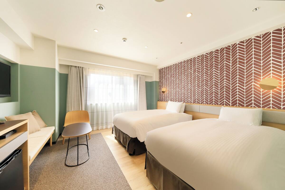 A hotel room with two beds, a desk, a chair, and a TV.