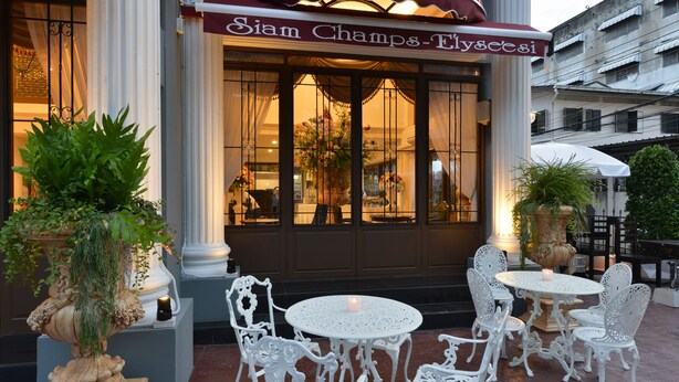 Uma área externa com assentos de ferro branco forjado, mesas, plantas em vasos e uma placa com a inscrição 'Siam Champs-Elysees'.
